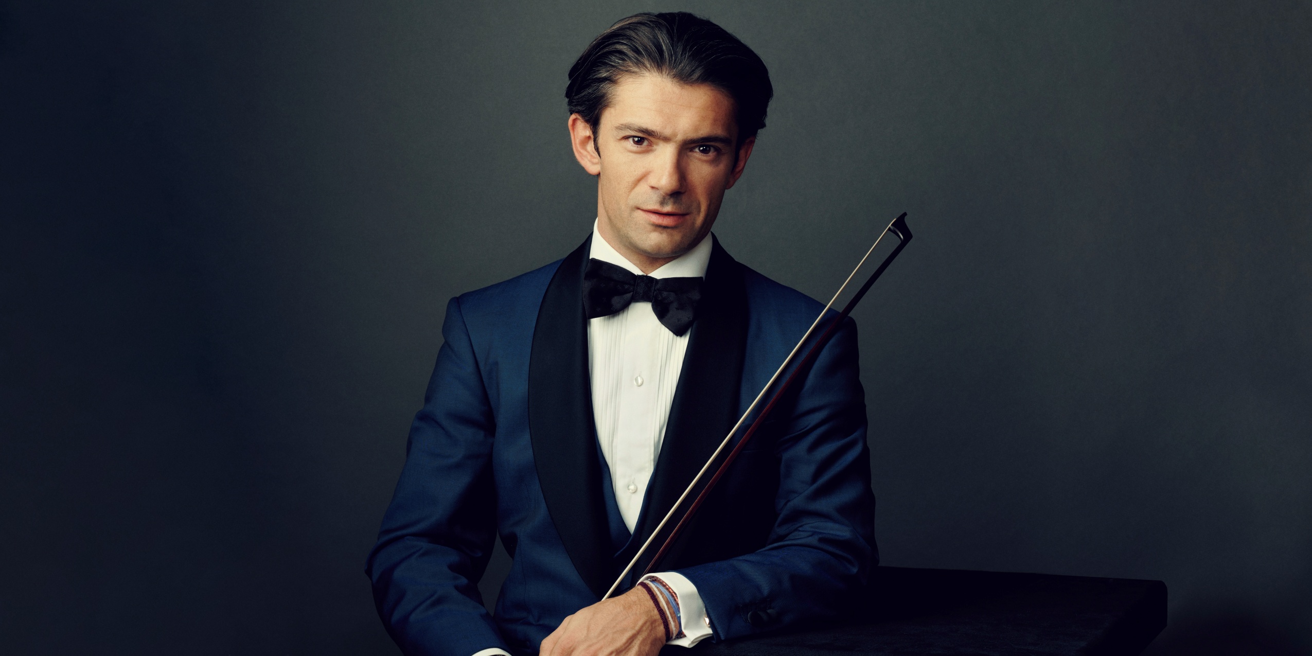 Mand med i blå blazer og sort butterfly, holder en cellobue foran mørkegrå baggrund.