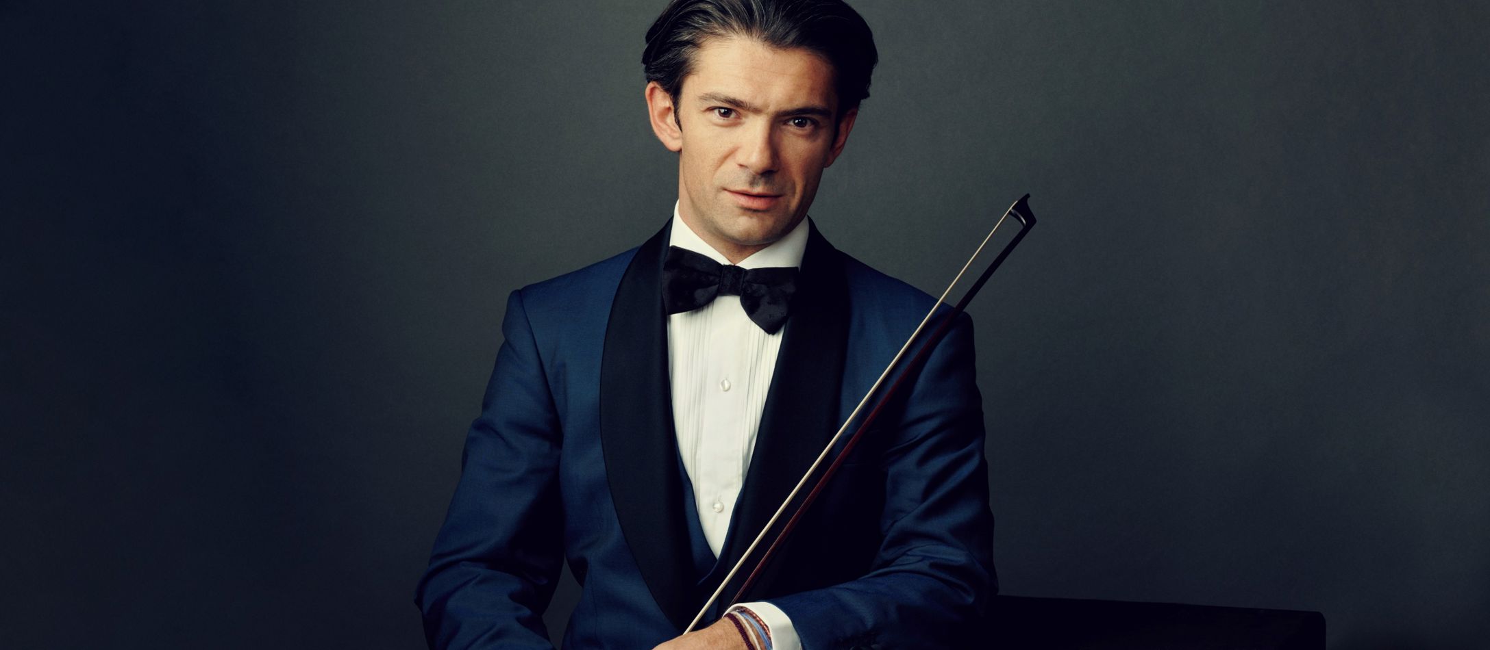 Mand med i blå blazer og sort butterfly, holder en cellobue foran mørkegrå baggrund.