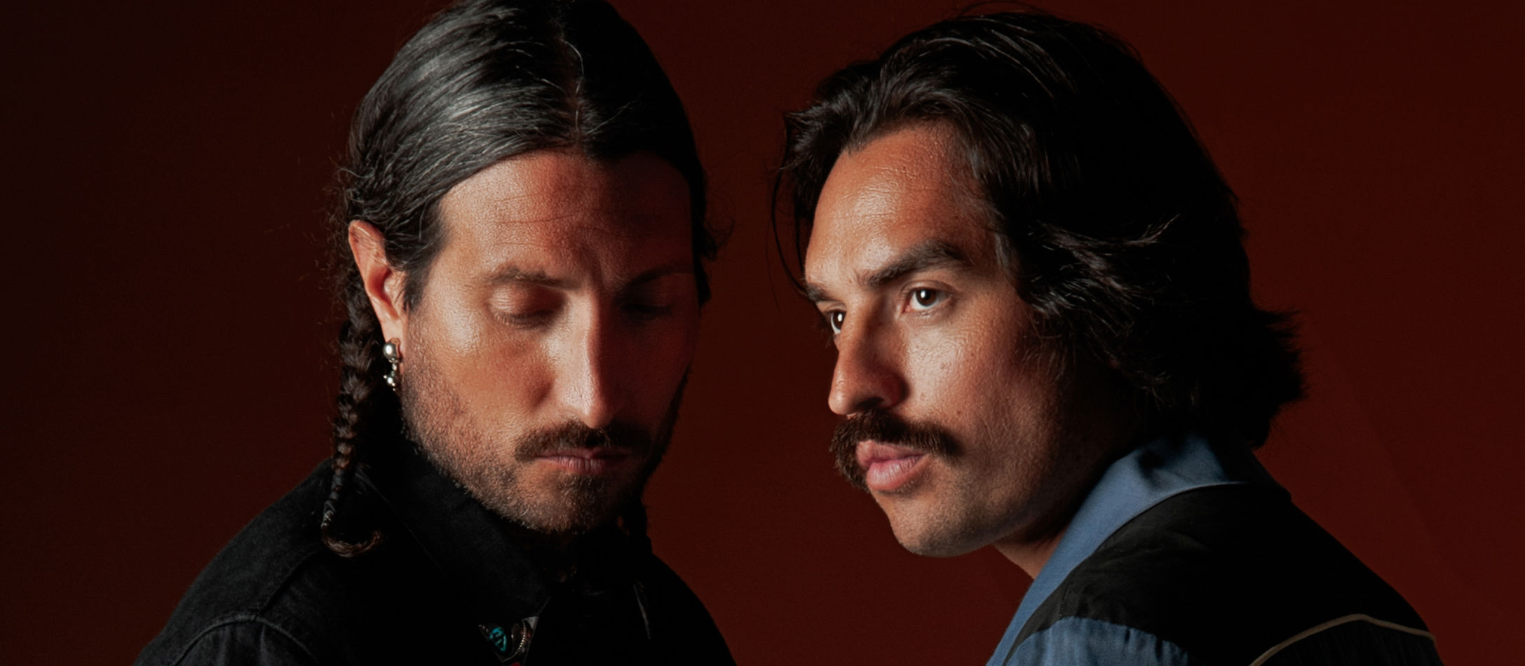 Hermanos Gutiérrez to brødre med alvorlige ansigtsudtryk iklædt elegant tøj foran mørkerød baggrund