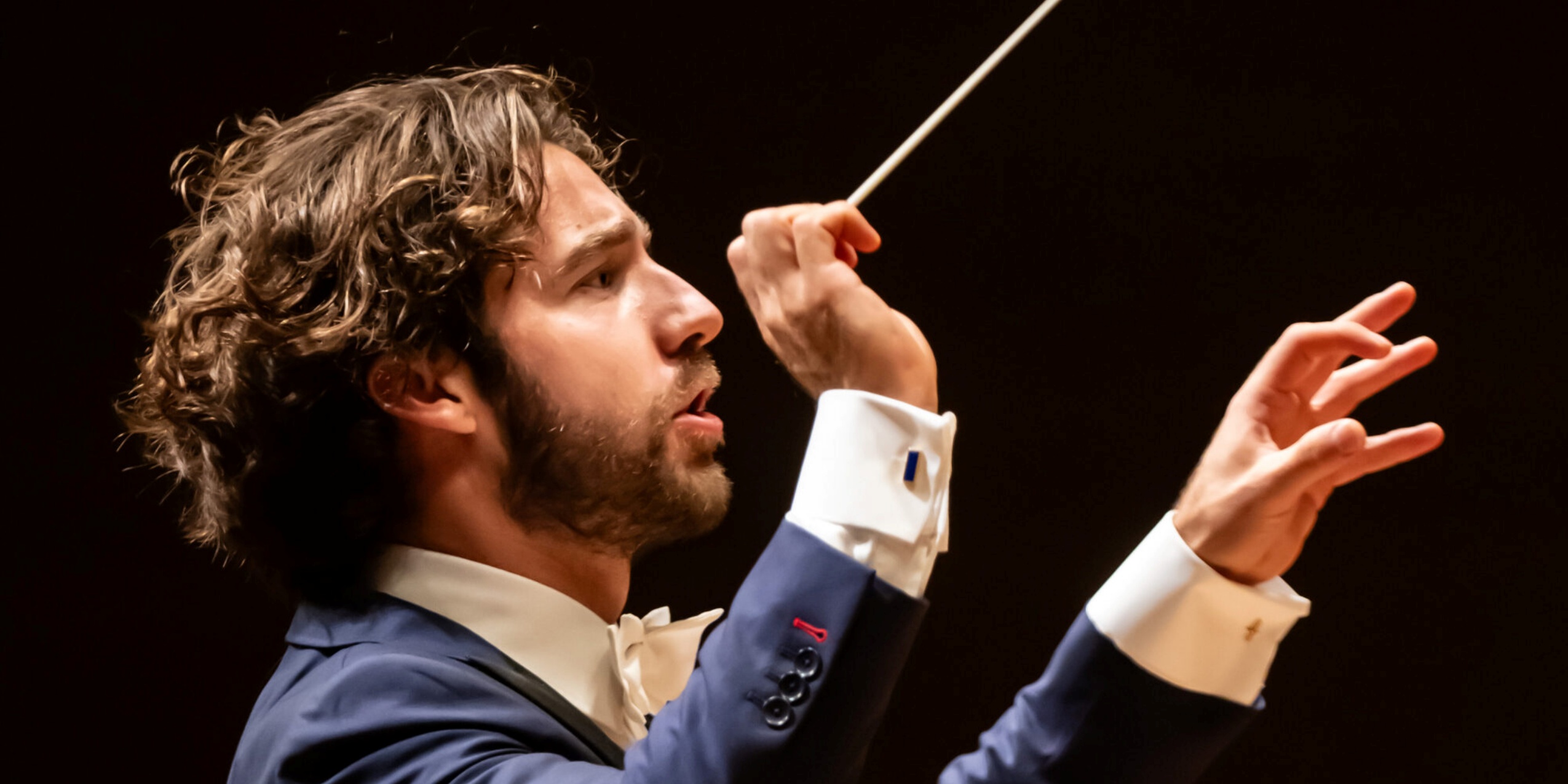 Mand med dirigentstok og armene løftet, i blå blazer og sort baggrund