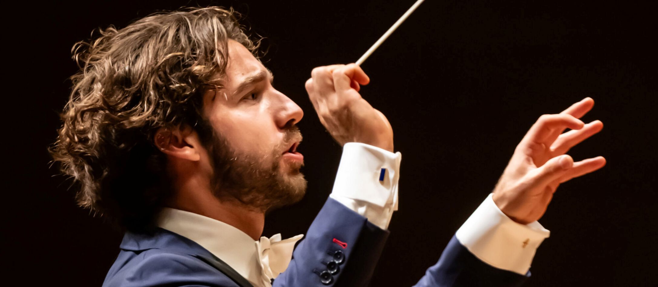 Mand med dirigentstok og armene løftet, i blå blazer og sort baggrund