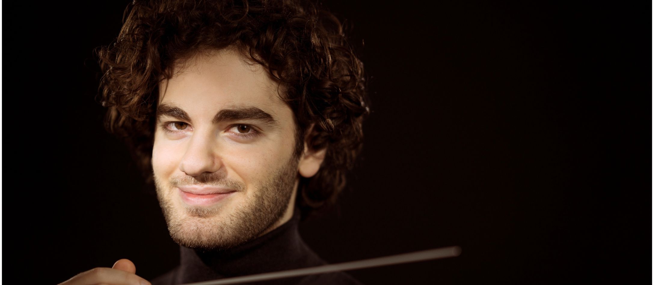 Mand med dirigentstok, smiler til kameraet.