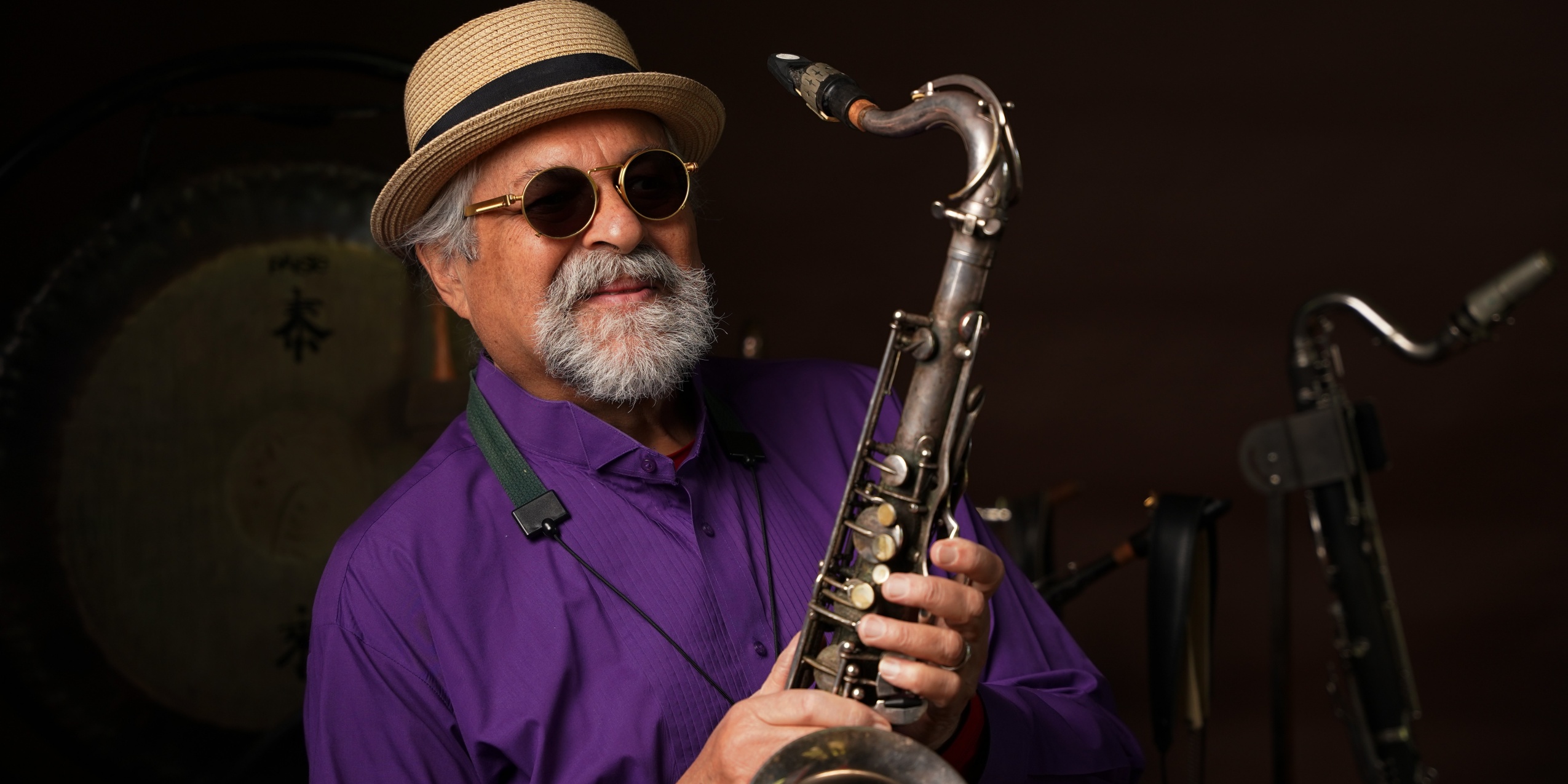 Mand med lilla skjorte, solbriller og stråhat, står med en saxofon.