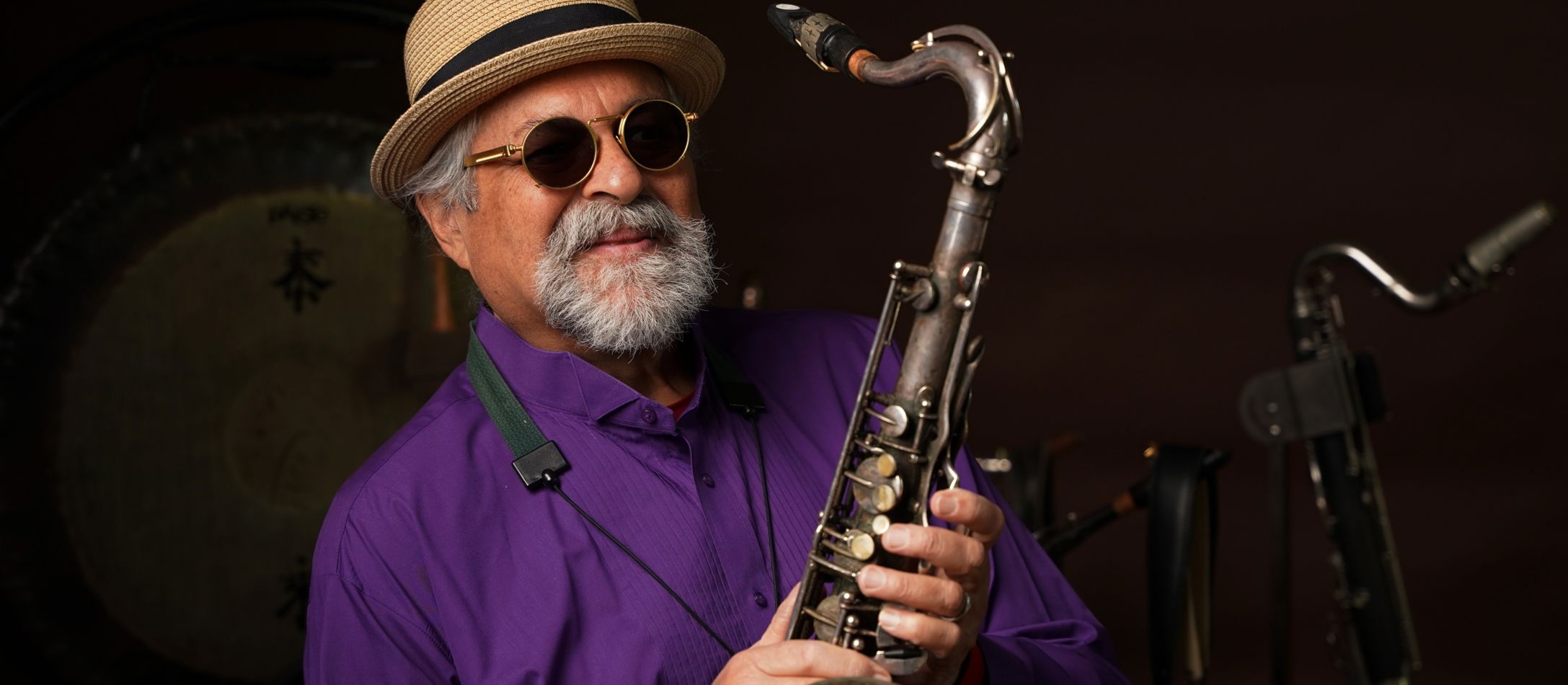 Mand med lilla skjorte, solbriller og stråhat, står med en saxofon.