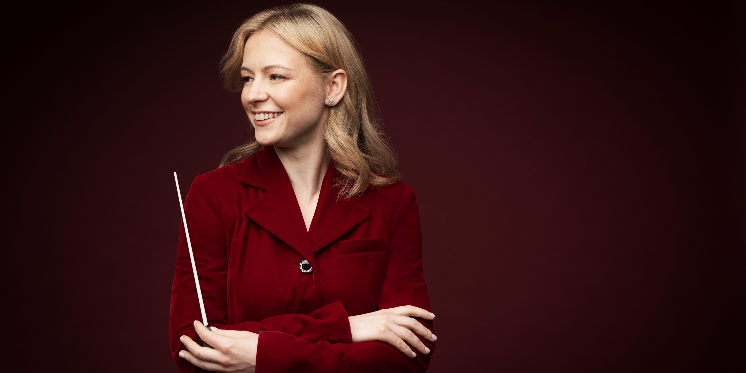 Kvinde smiler i rød jakke med en dirigentstok, foran mørkerød væg.