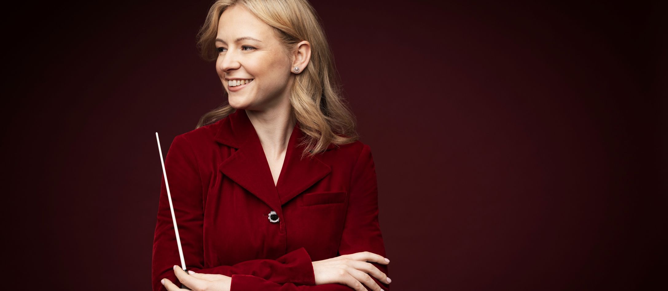 Kvinde smiler i rød jakke med en dirigentstok, foran mørkerød væg.