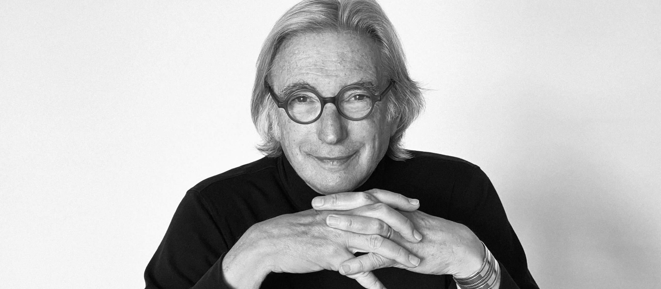 Med sine runde briller på samler smilendeMichael Tilson Thomas fingrene under hagen med albuerne hvilende på bordet foran.