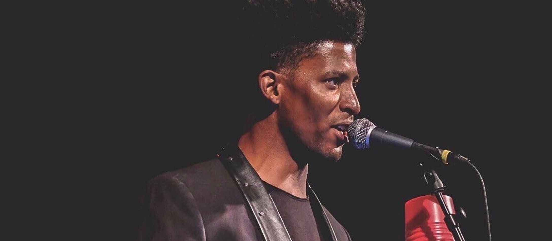 Henri Benitez i profil med glinsende hud og højt mørkt afrohår med rød percussioninstrument i hånden taler ind i mikrofon.