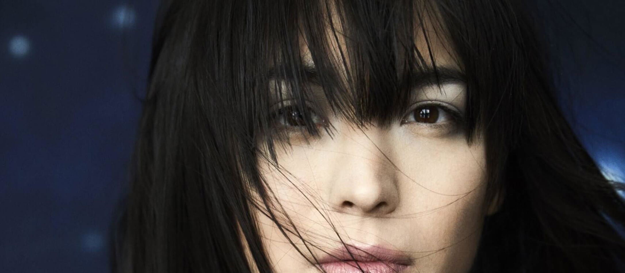 Alice Sara Ott med mørkt pandehår, der falder foran dybe, brune øjne med bare skuldre og høj hals på bluse med blå baggrund