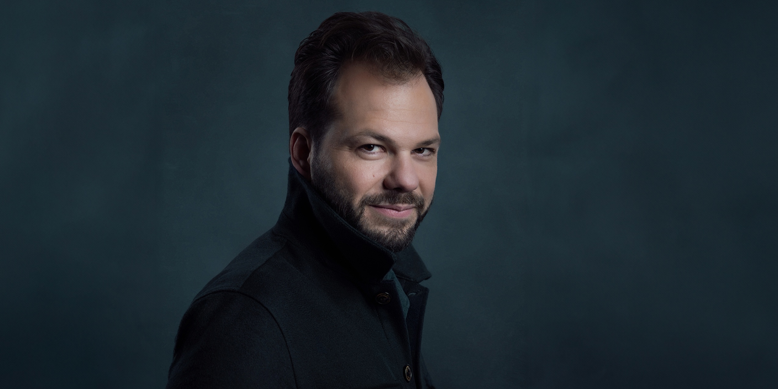 Man i mørk jakke med opslået krave foran mørkeblå væg.