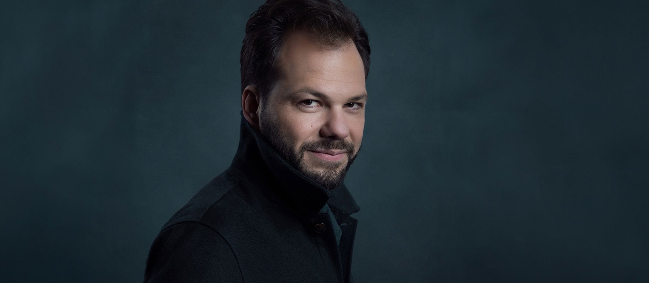 Man i mørk jakke med opslået krave foran mørkeblå væg.