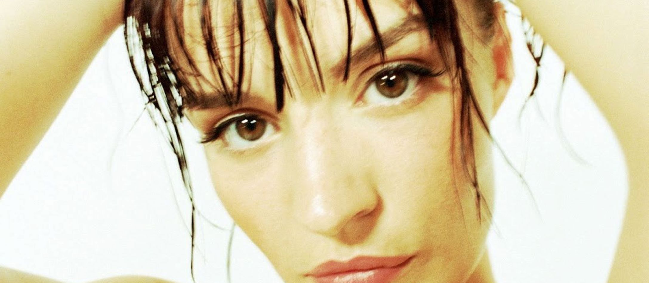 Thea Dora med fængslende brune øjne, mørkt pandehår og hænderne i opsat hår på hvid baggrund