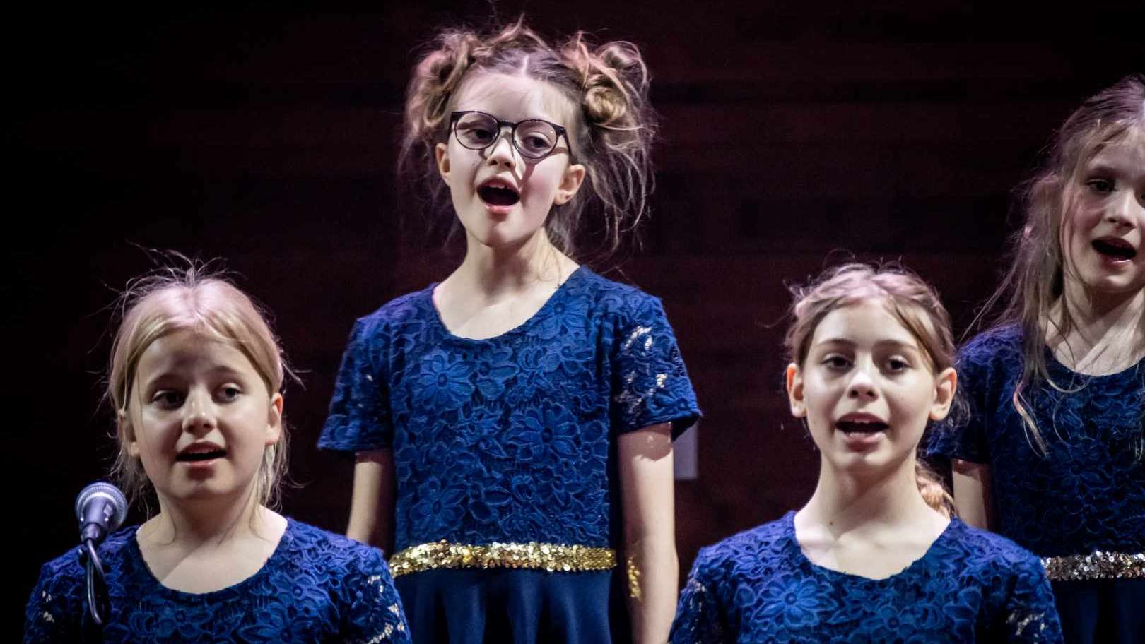 Fire glade børn fra DR Børnekoret iført blå blondekjoler synger foran sort baggrund