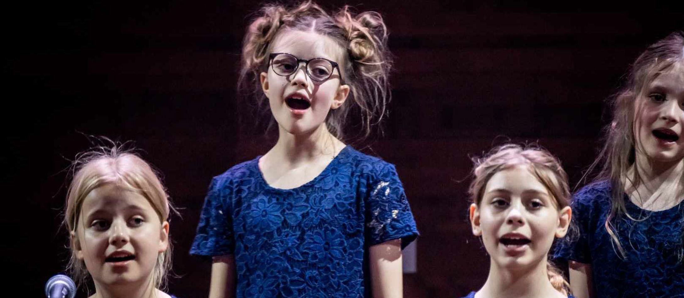 Fire glade børn fra DR Børnekoret iført blå blondekjoler synger foran sort baggrund