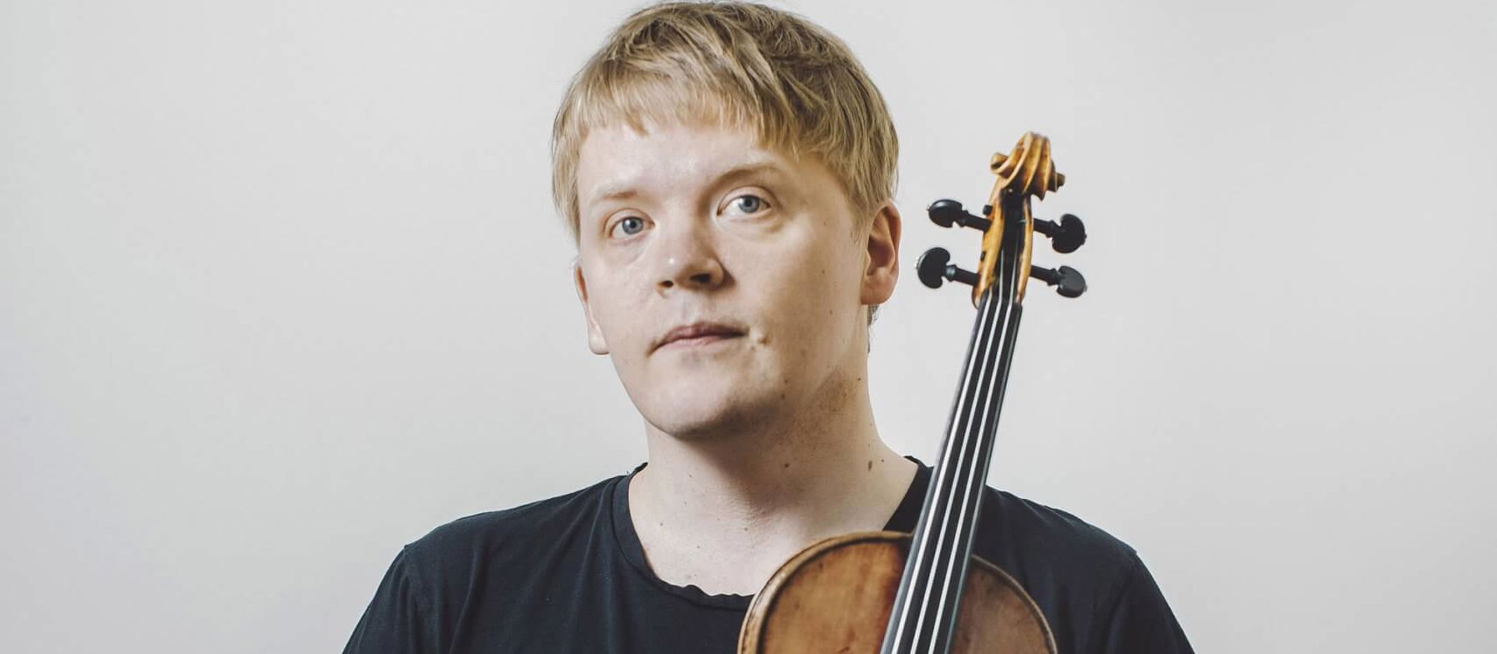 Pekka Kuusisto iført sort t-shirt og sorte bukser foran hvid væg holder sin fine violin i den ene hånd den anden i lommen.
