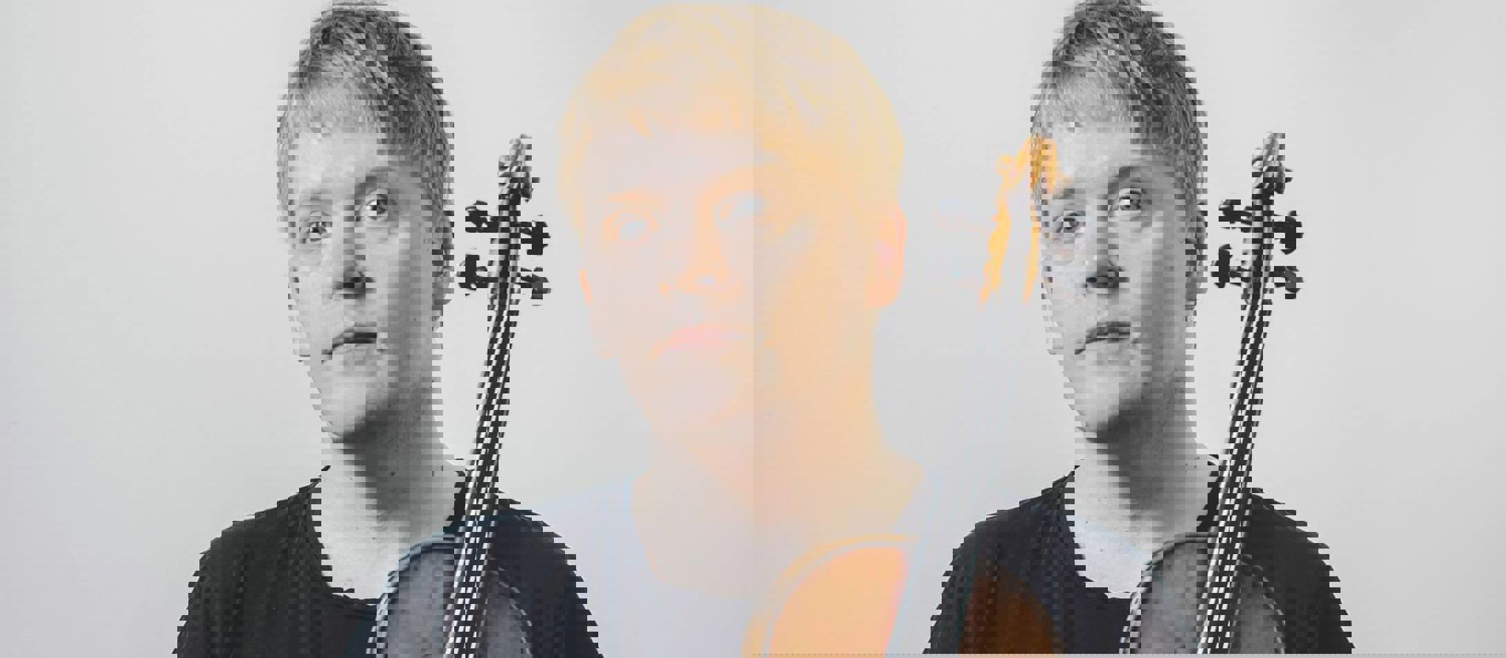 Pekka Kuusisto iført sort t-shirt og sorte bukser foran hvid væg holder sin fine violin i den ene hånd den anden i lommen.