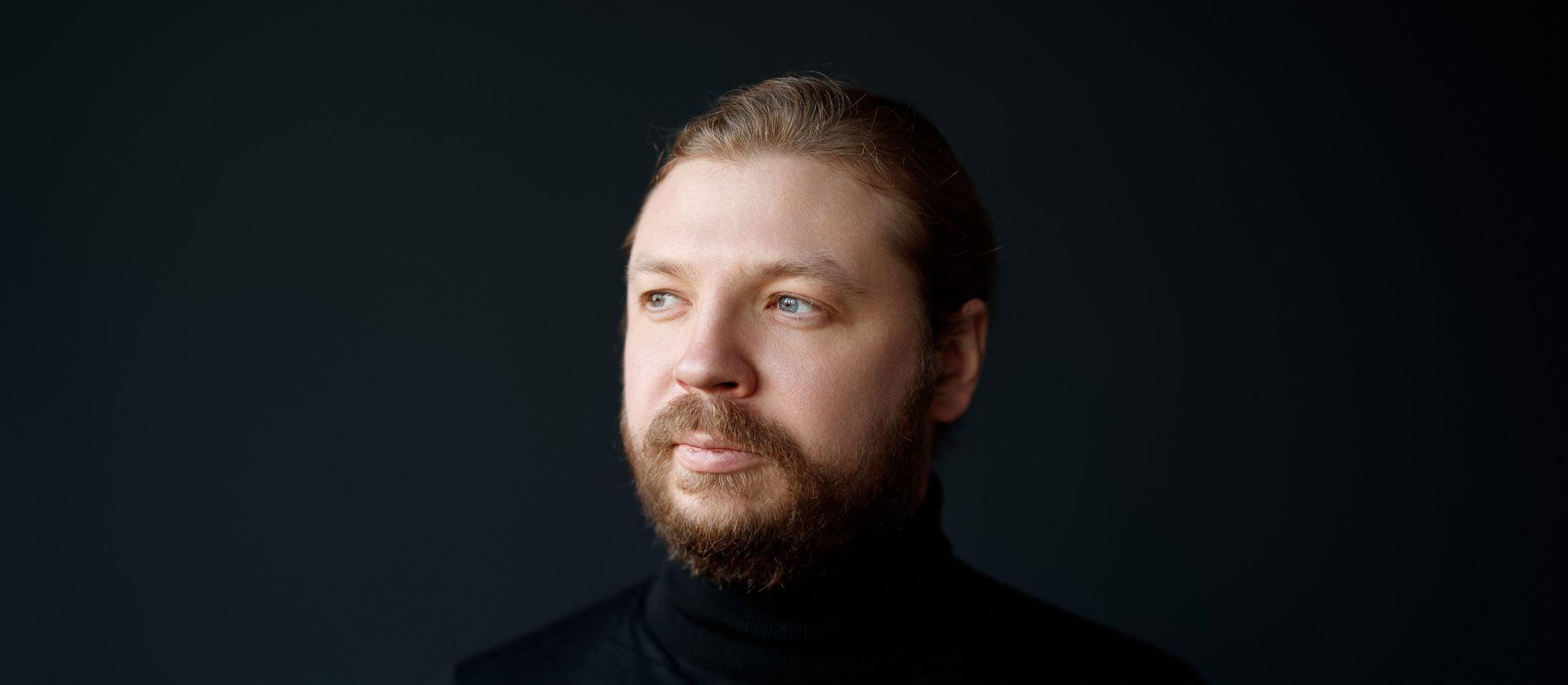 Pianisten Denis Kozhukhin med sort halskrave oh rødligt hår og skæg og blå øjne kigger mod sin højre foran sort baggrund.