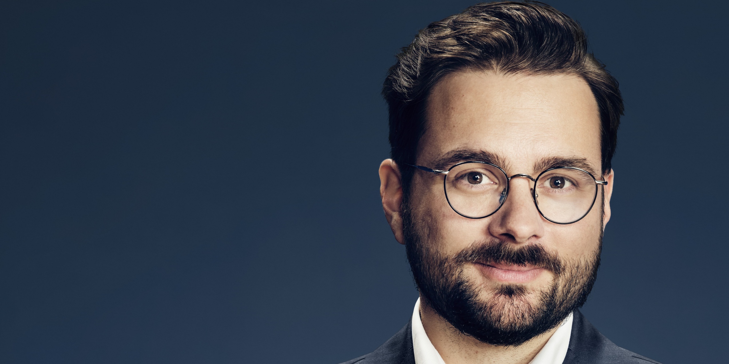 Mand med briller kigger ind i kameraet, foran mørk gråblå væg.