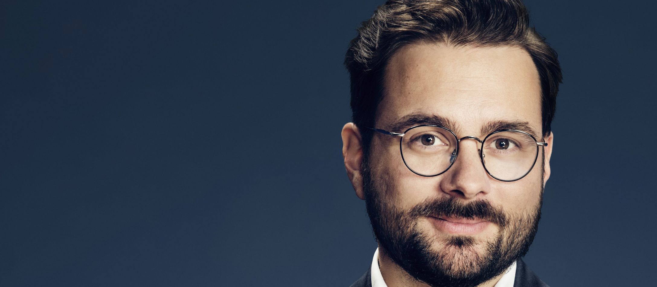 Mand med briller kigger ind i kameraet, foran mørk gråblå væg.