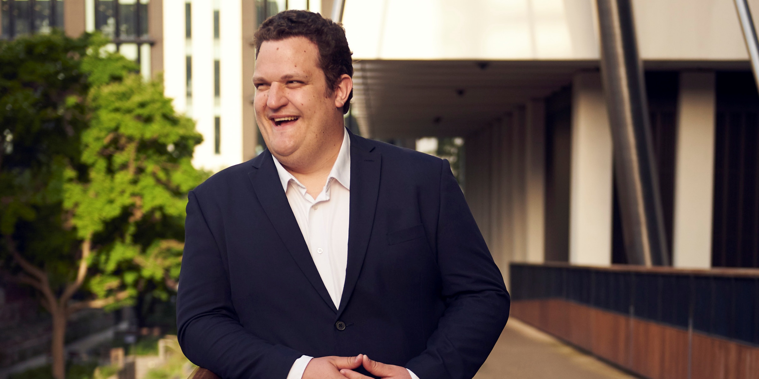 Mand der smiler og læner sig op ad et rækværk på en bro ved en bygning