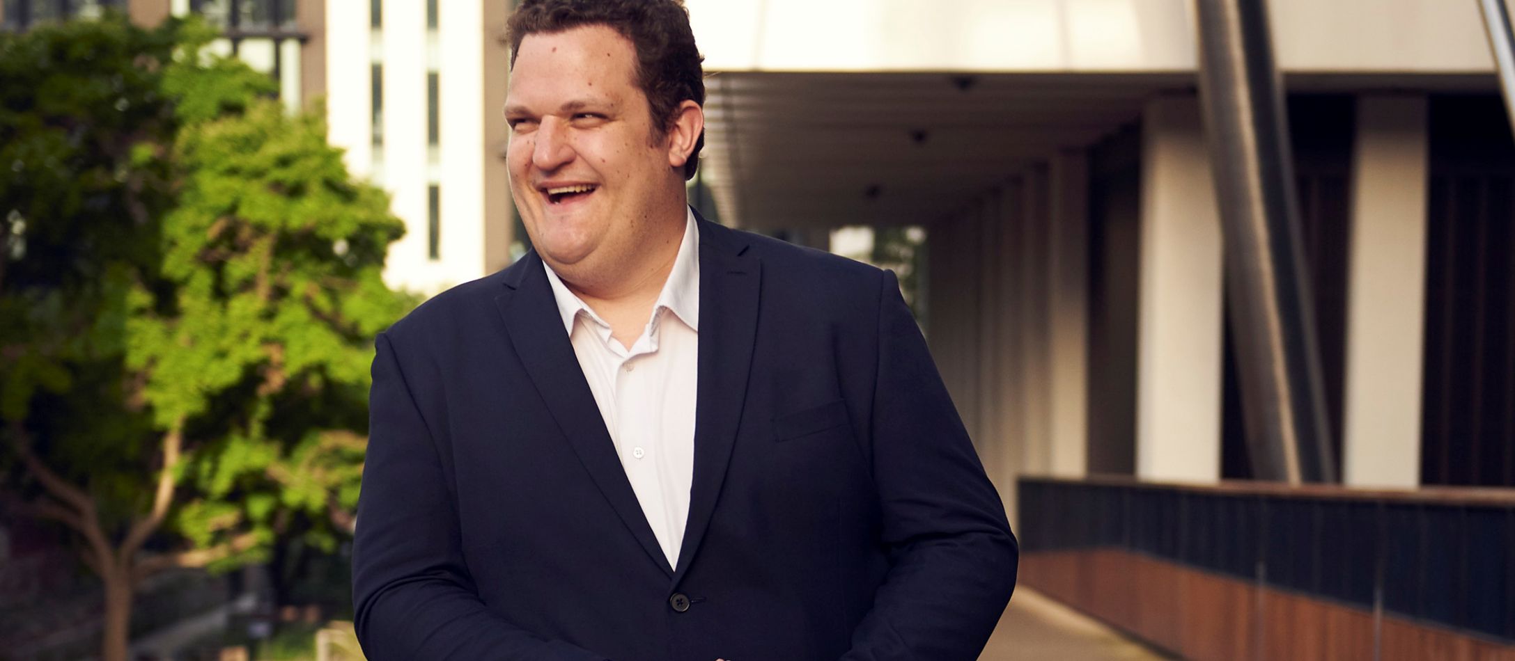 Mand der smiler og læner sig op ad et rækværk på en bro ved en bygning