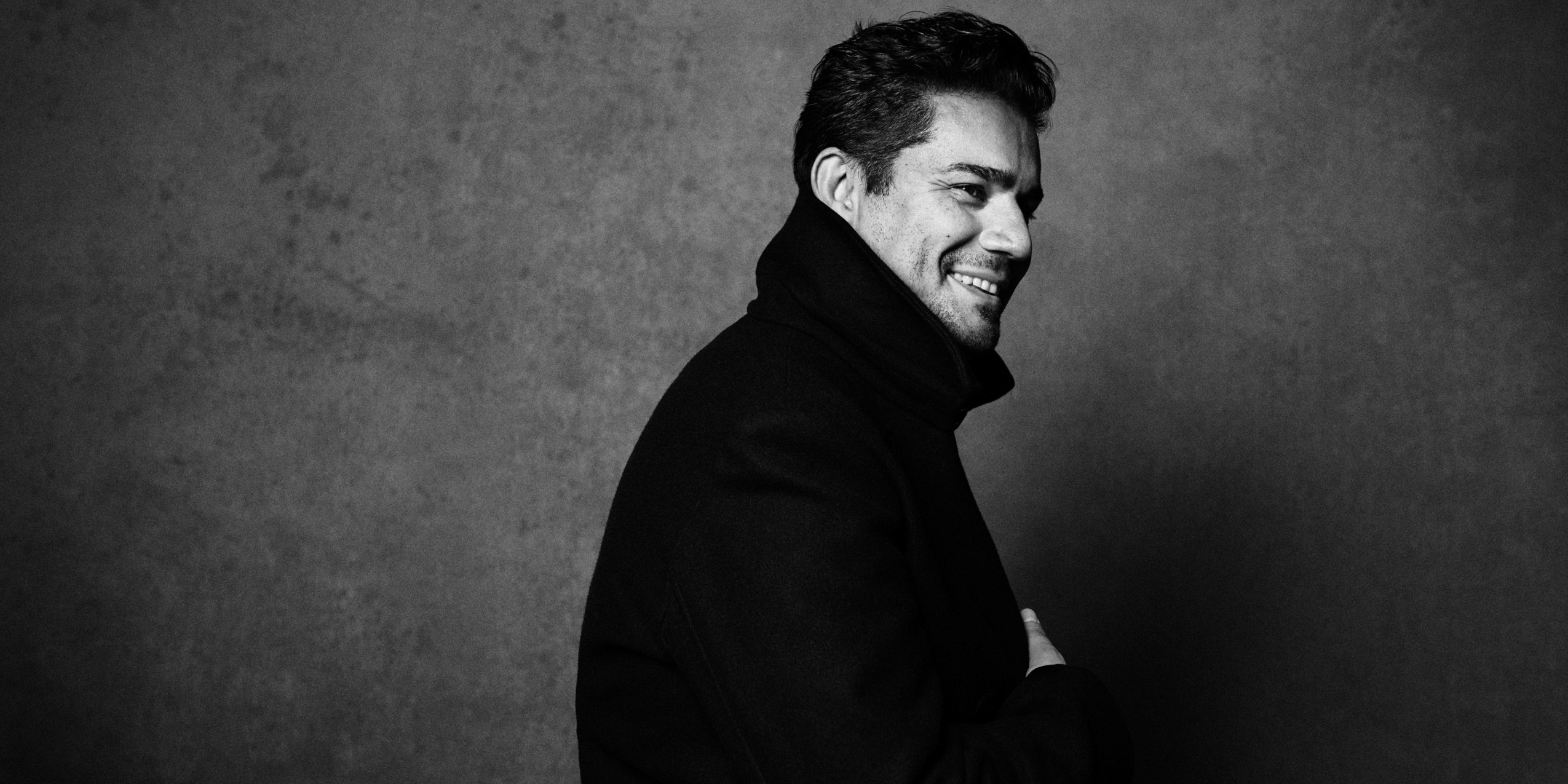 Mand i sort frakke smiler foran grå væg