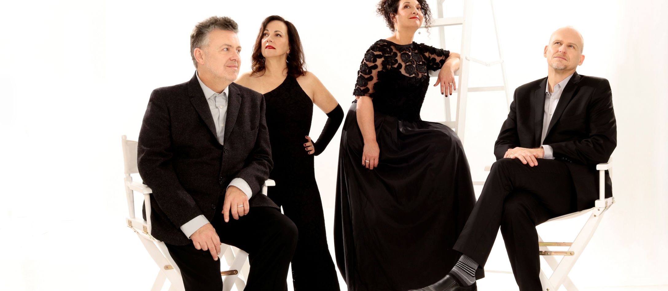 Fire sangere i pænt sort tøj, stå og sidder ved to hvide stole og en hvid stige.