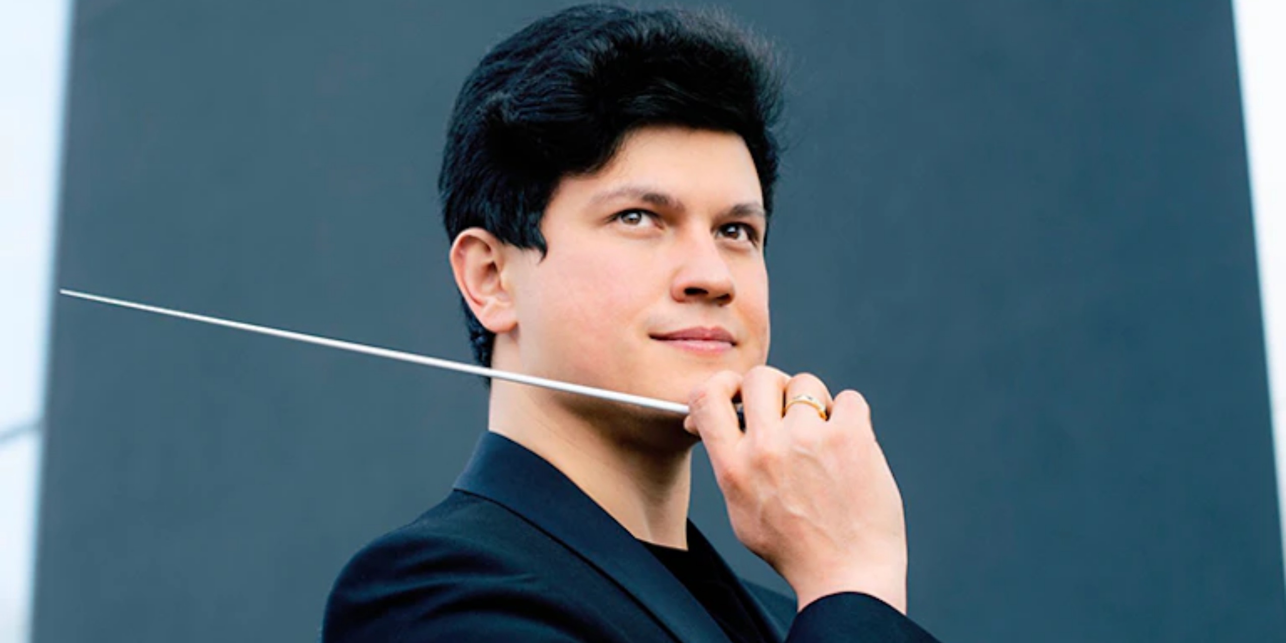 Mand med dirigentstok og hånden løftet til ansigtet, i sort blazer og grå baggrund