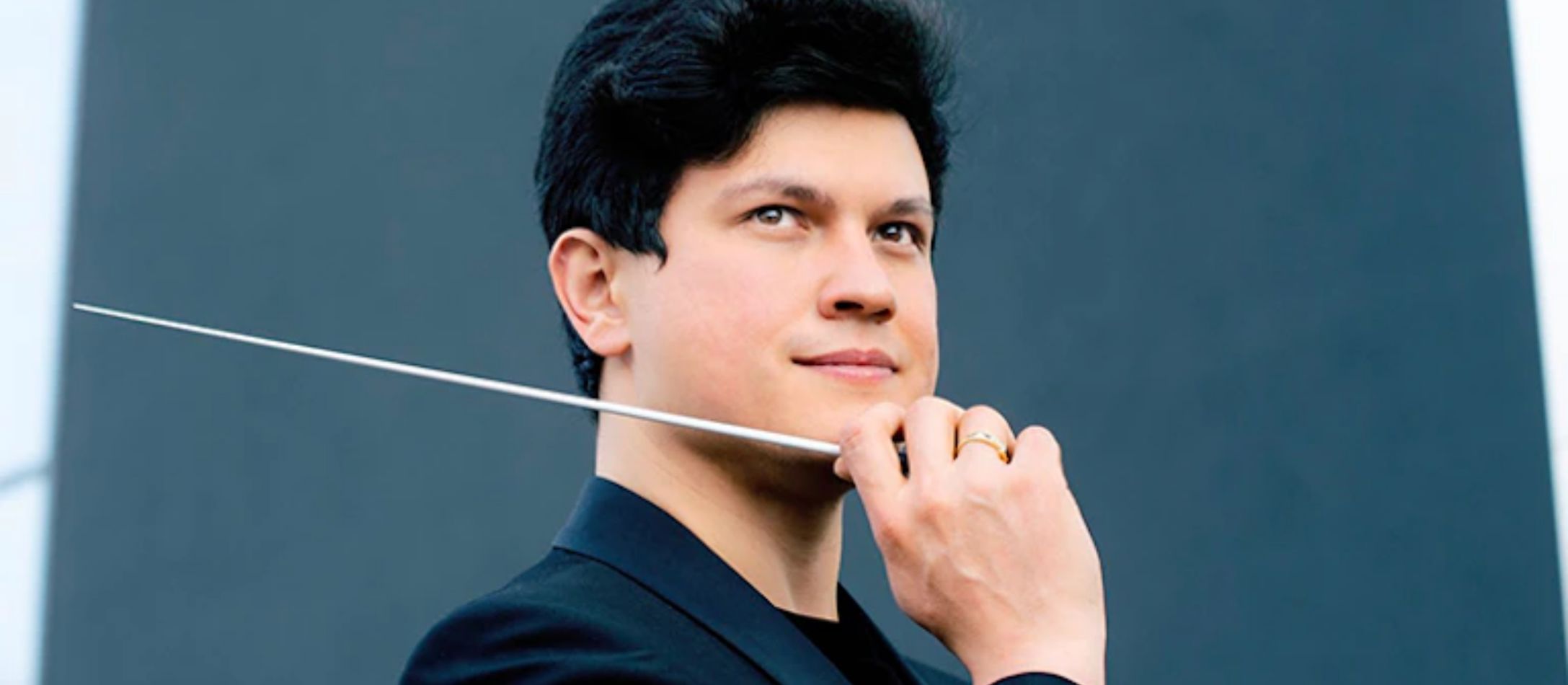 Mand med dirigentstok og hånden løftet til ansigtet, i sort blazer og grå baggrund