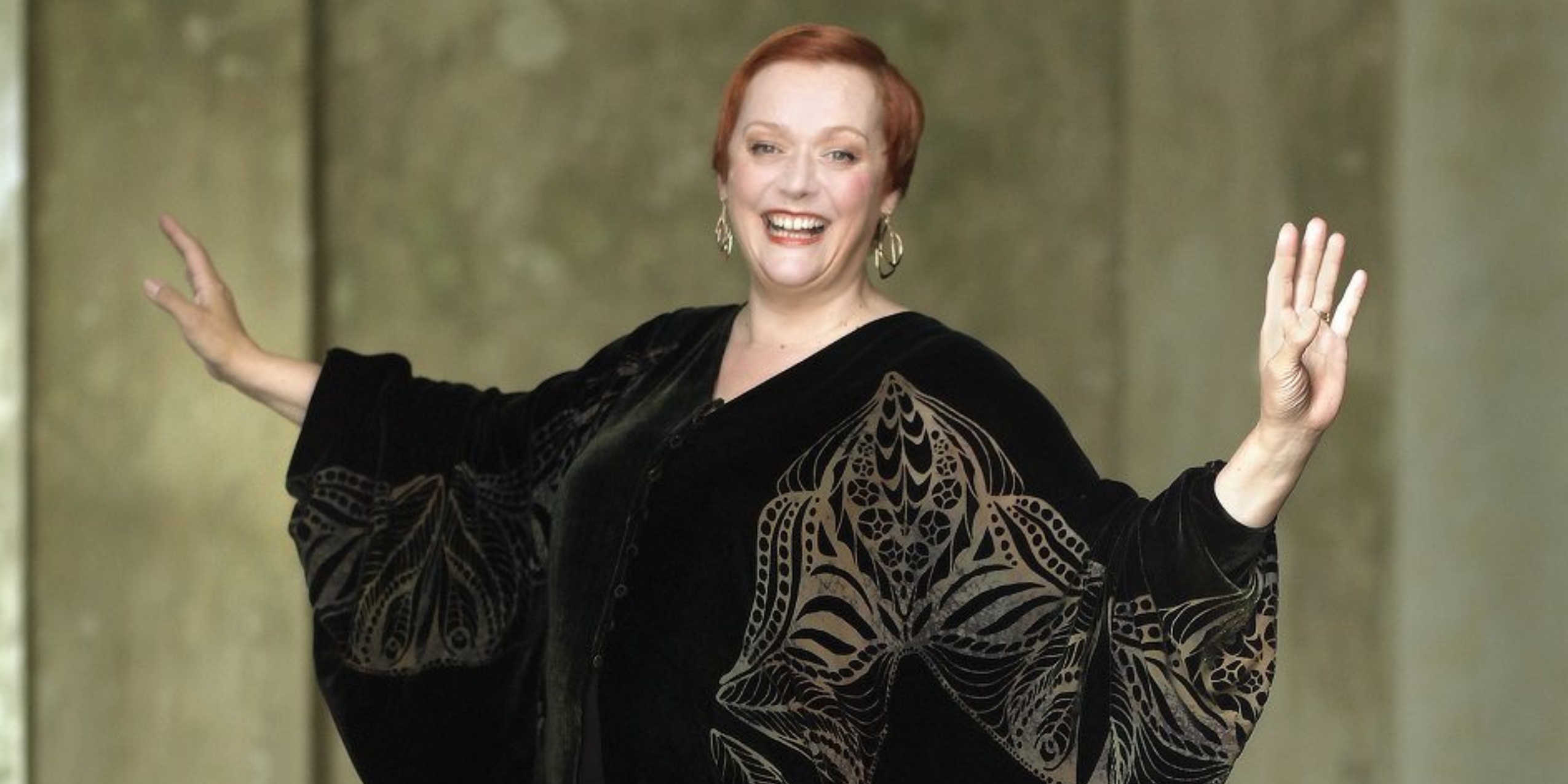 Kvinde i sort kjole med guld broderi, står smilende med armene ud til hver side.