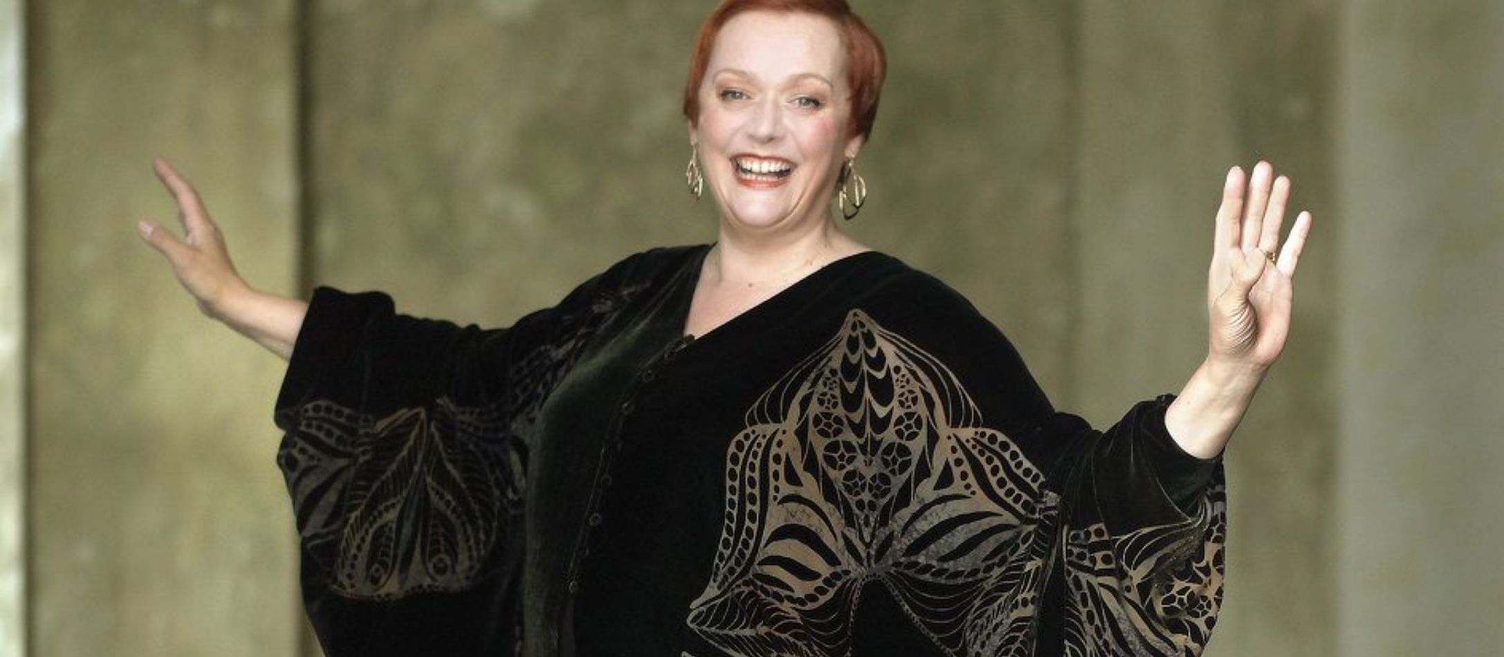 Kvinde i sort kjole med guld broderi, står smilende med armene ud til hver side.