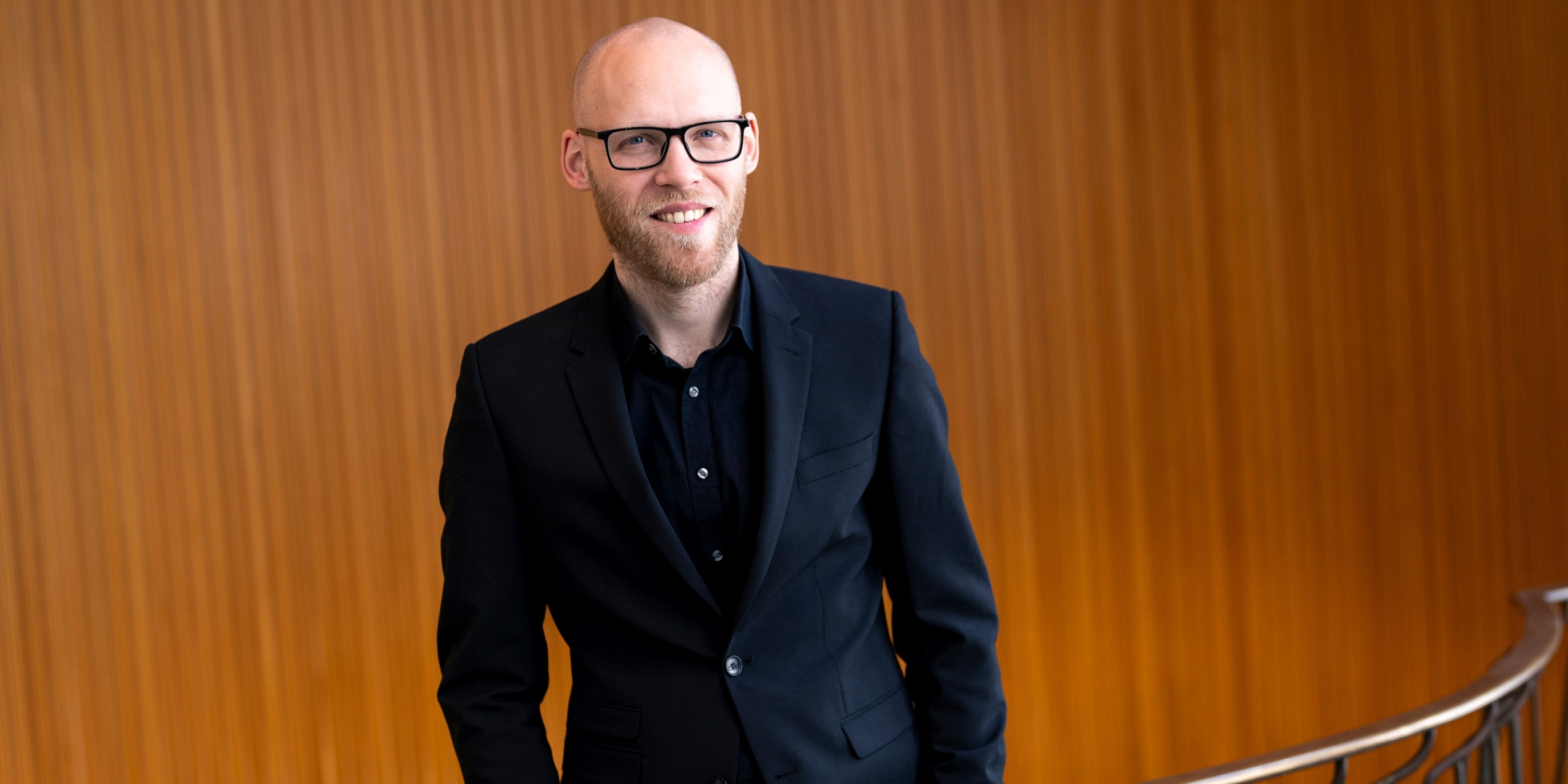 Mand med briller i sort tøj, foran et gelænder og træ væg.