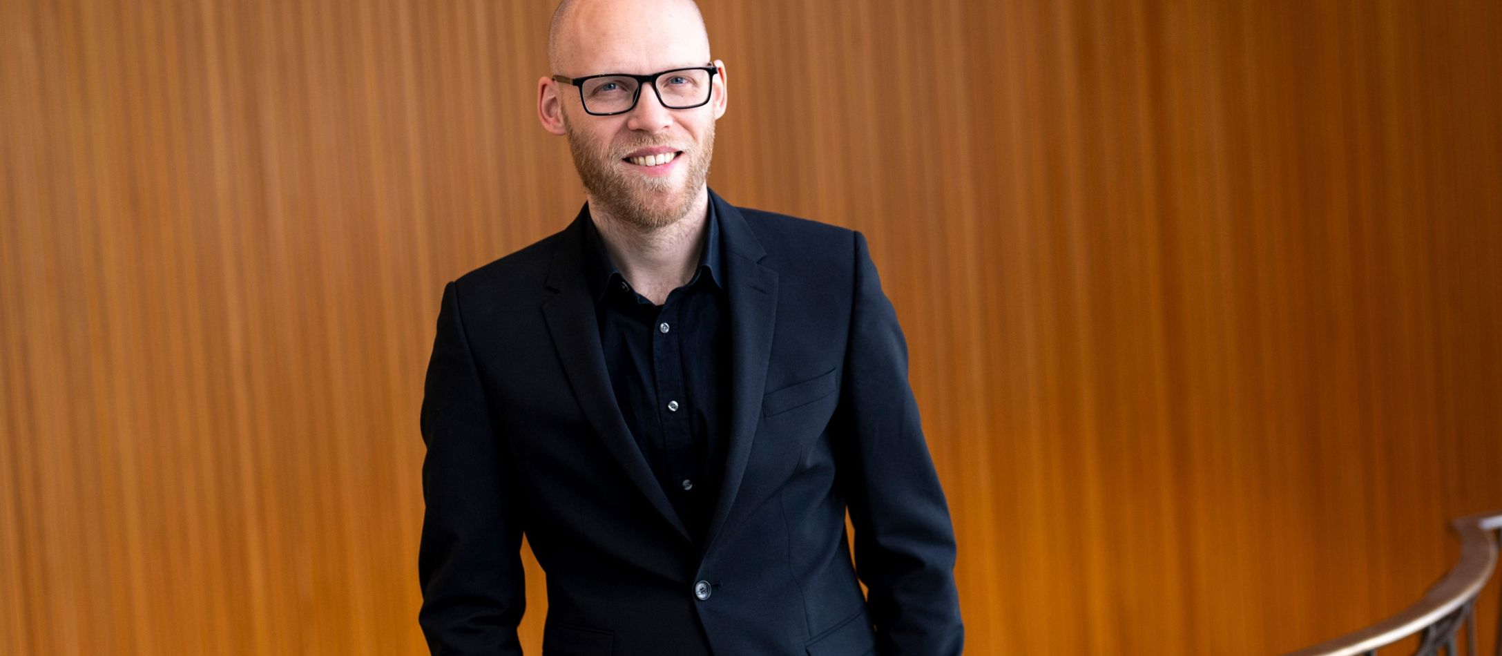 Mand med briller i sort tøj, foran et gelænder og træ væg.