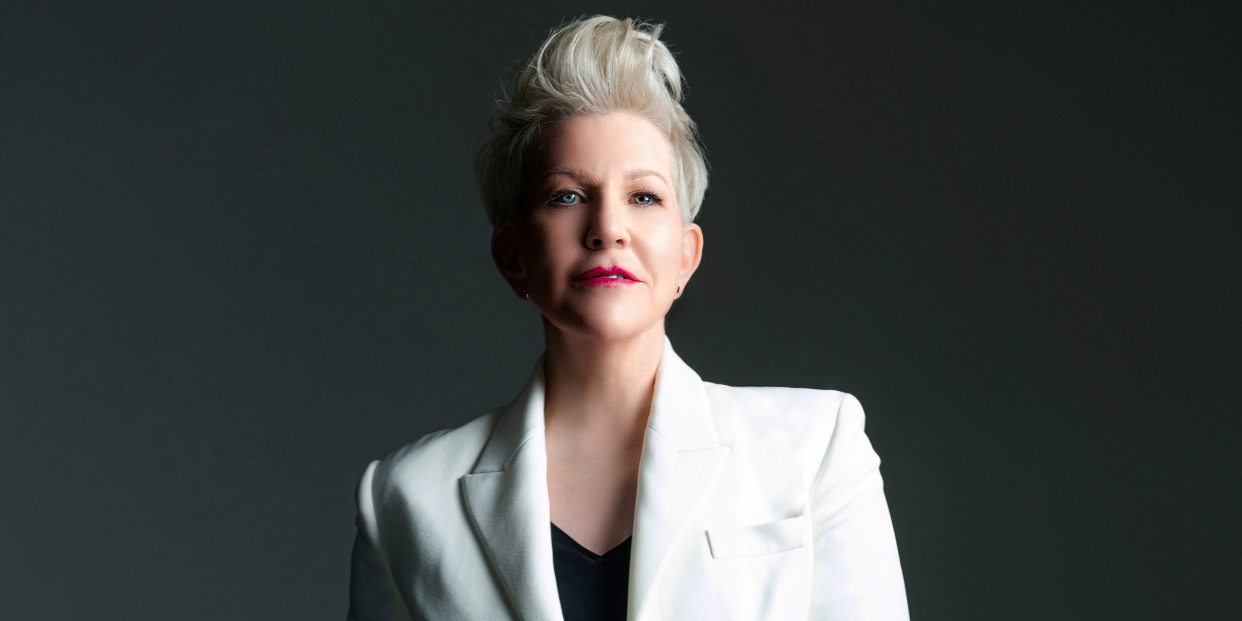 Kvinde i hvid blazer, kigger ind i kameraet foran en mørkegrå baggrund.