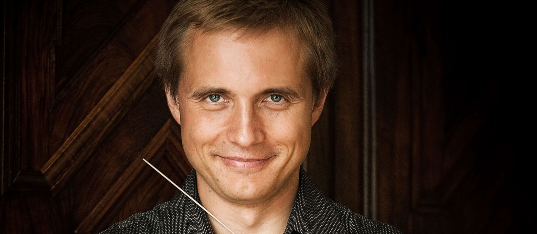 Dirigenten Vasily Petrenko med korslagte arme smiler behageligt med dirigentstang i højre hånd ført op mod ansigtet.