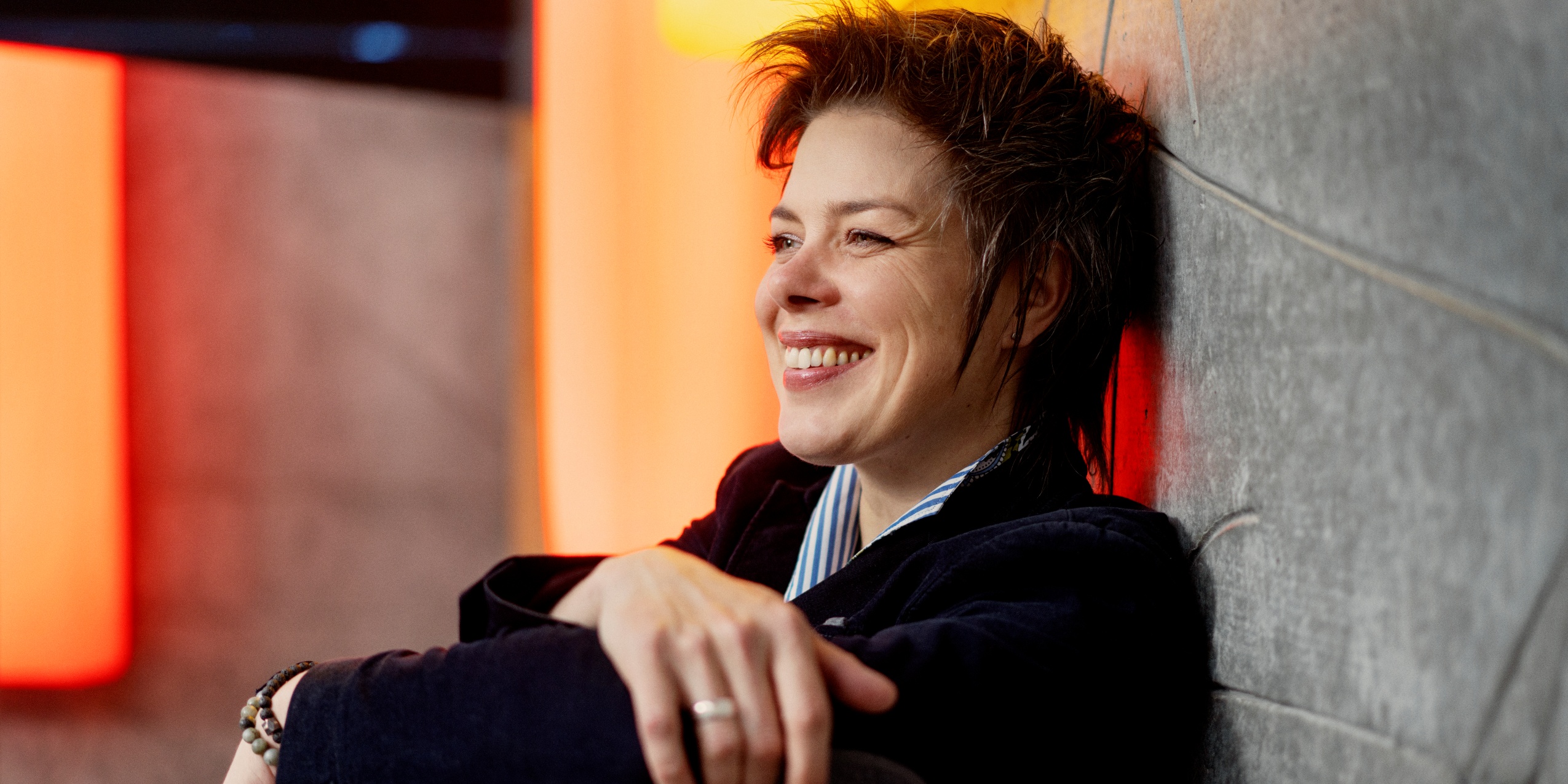 Kvinde mørkeblå jakke og blåstribet skjorte, sidder smilende op ad en betonvæg.