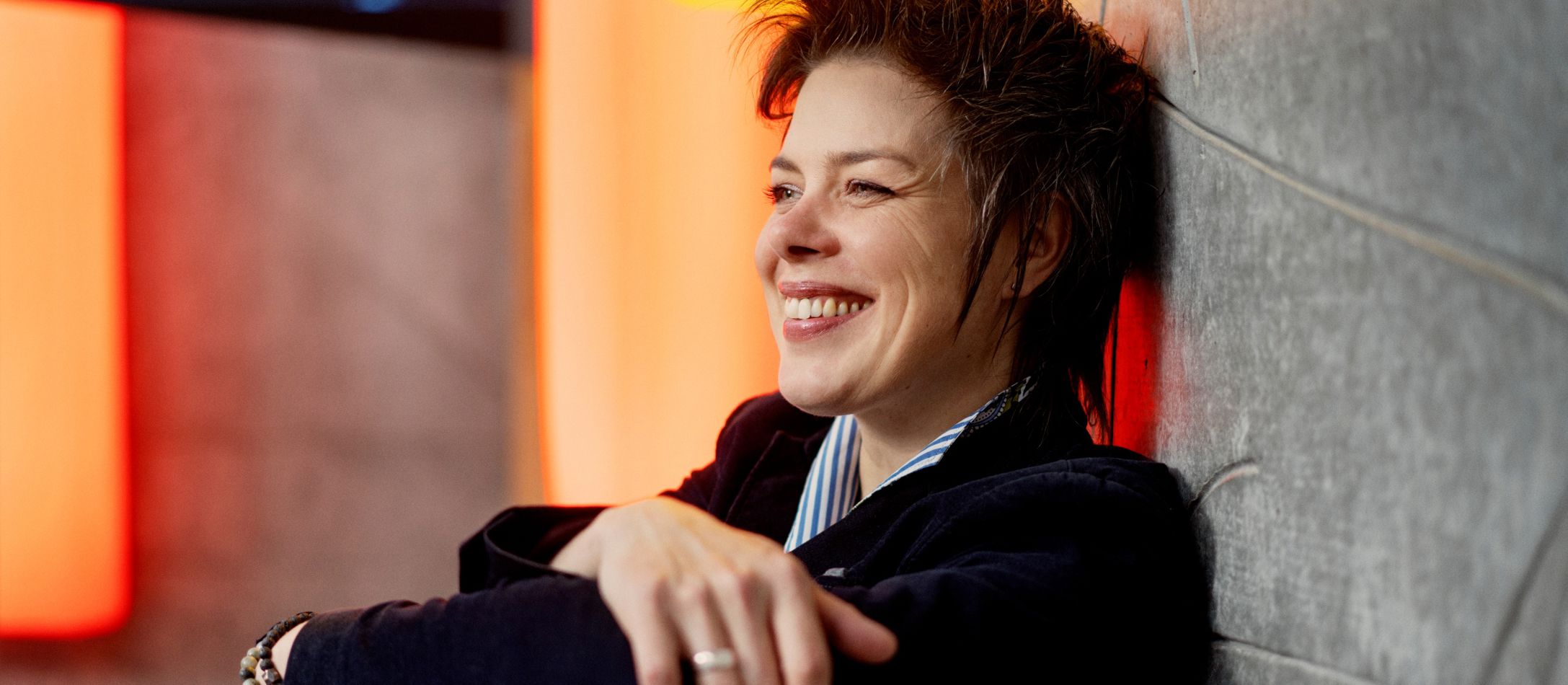 Kvinde mørkeblå jakke og blåstribet skjorte, sidder smilende op ad en betonvæg.