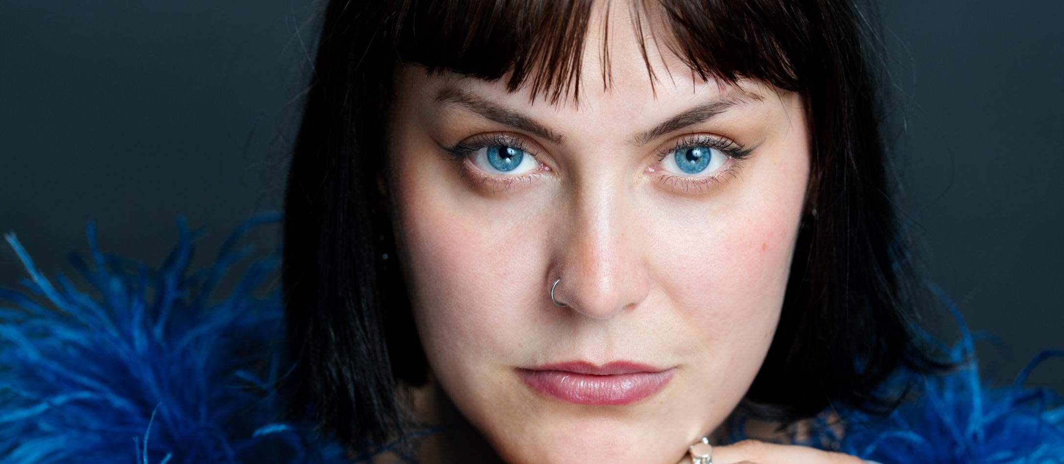 Kvinde med blå øjne og blå fjerboa, kigger ind i kameraet.