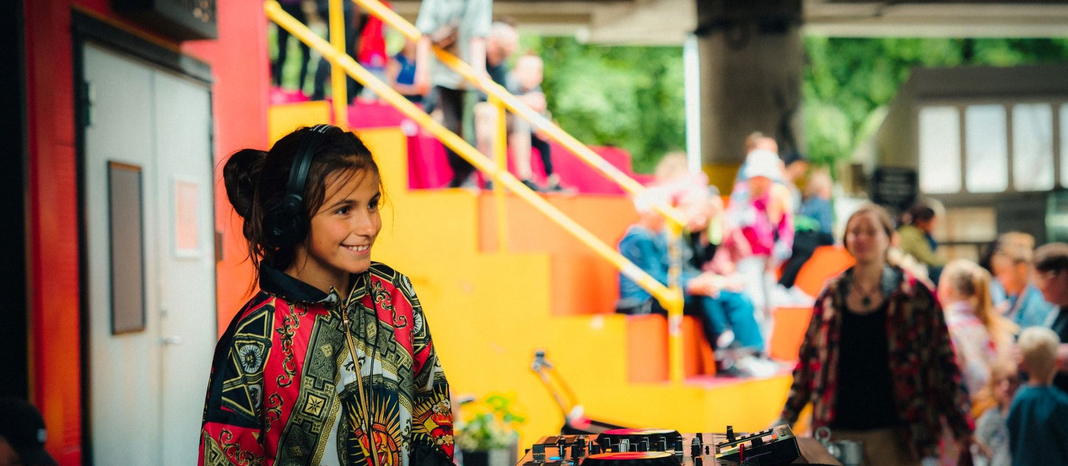 En ung DJ med farverig jakke på iført hovedtelefoner foran en dj-pult og mennesker i baggrunden