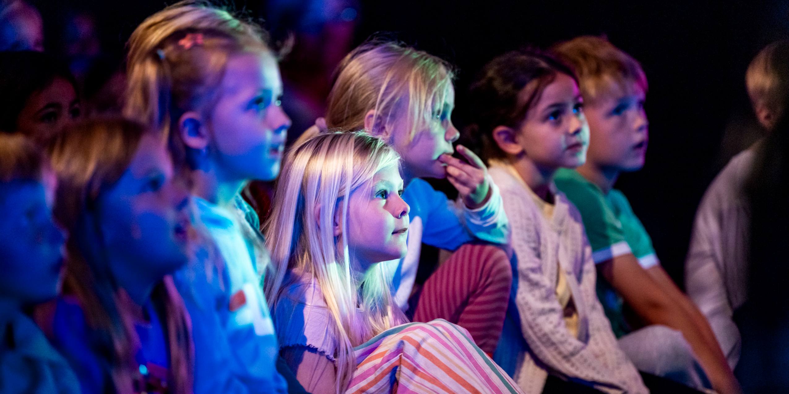 Børn til skolekoncert sidder i blåt skær og kigger på scenen