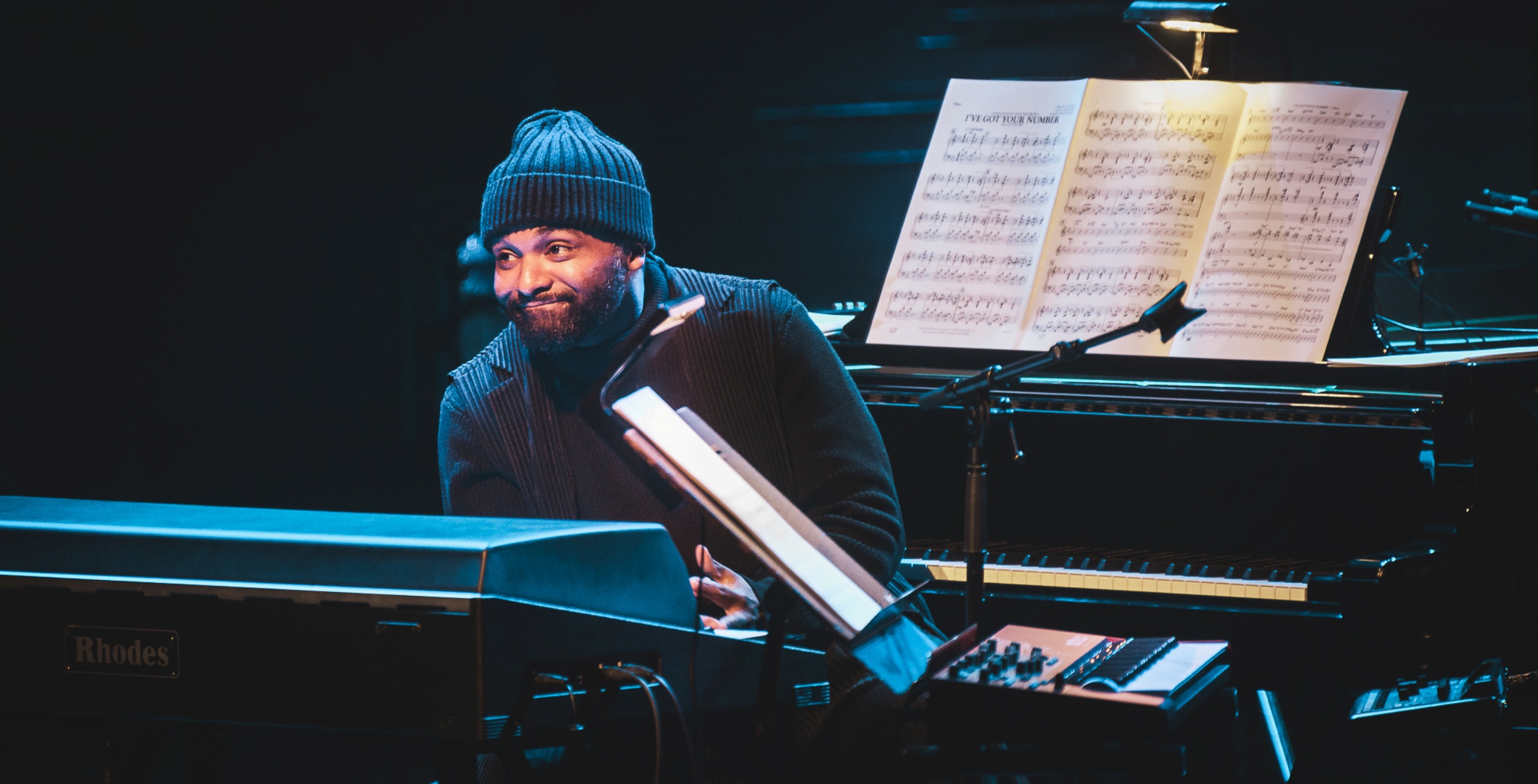 Mand i sort og med hue på, sidder mellem to klaverer og smiler.