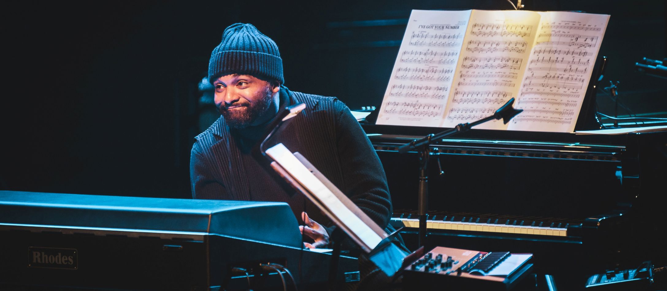 Mand i sort og med hue på, sidder mellem to klaverer og smiler.