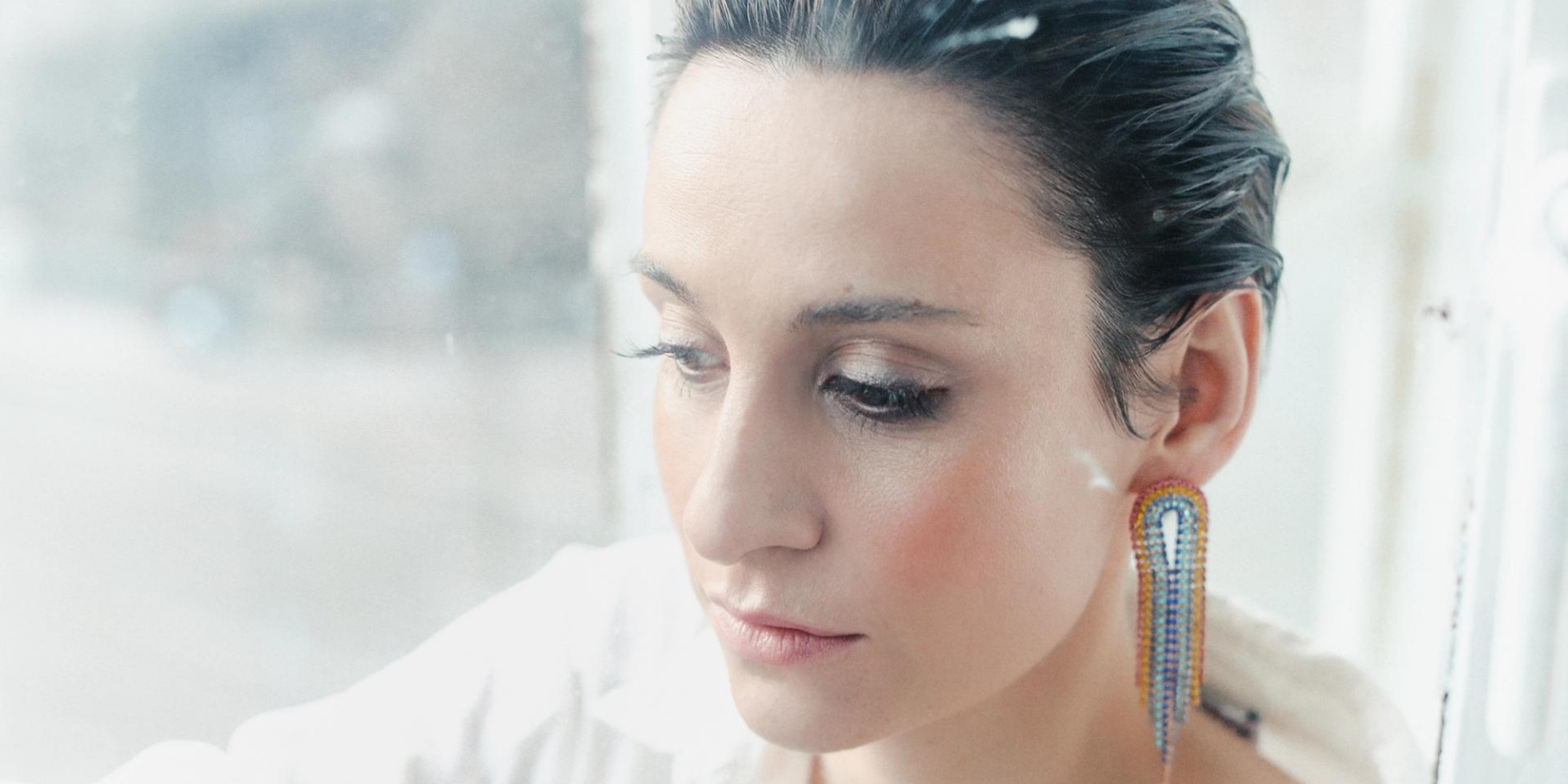 Kvinde i hvidt med en regnbueørering foran et vindue med hvide gardiner. 