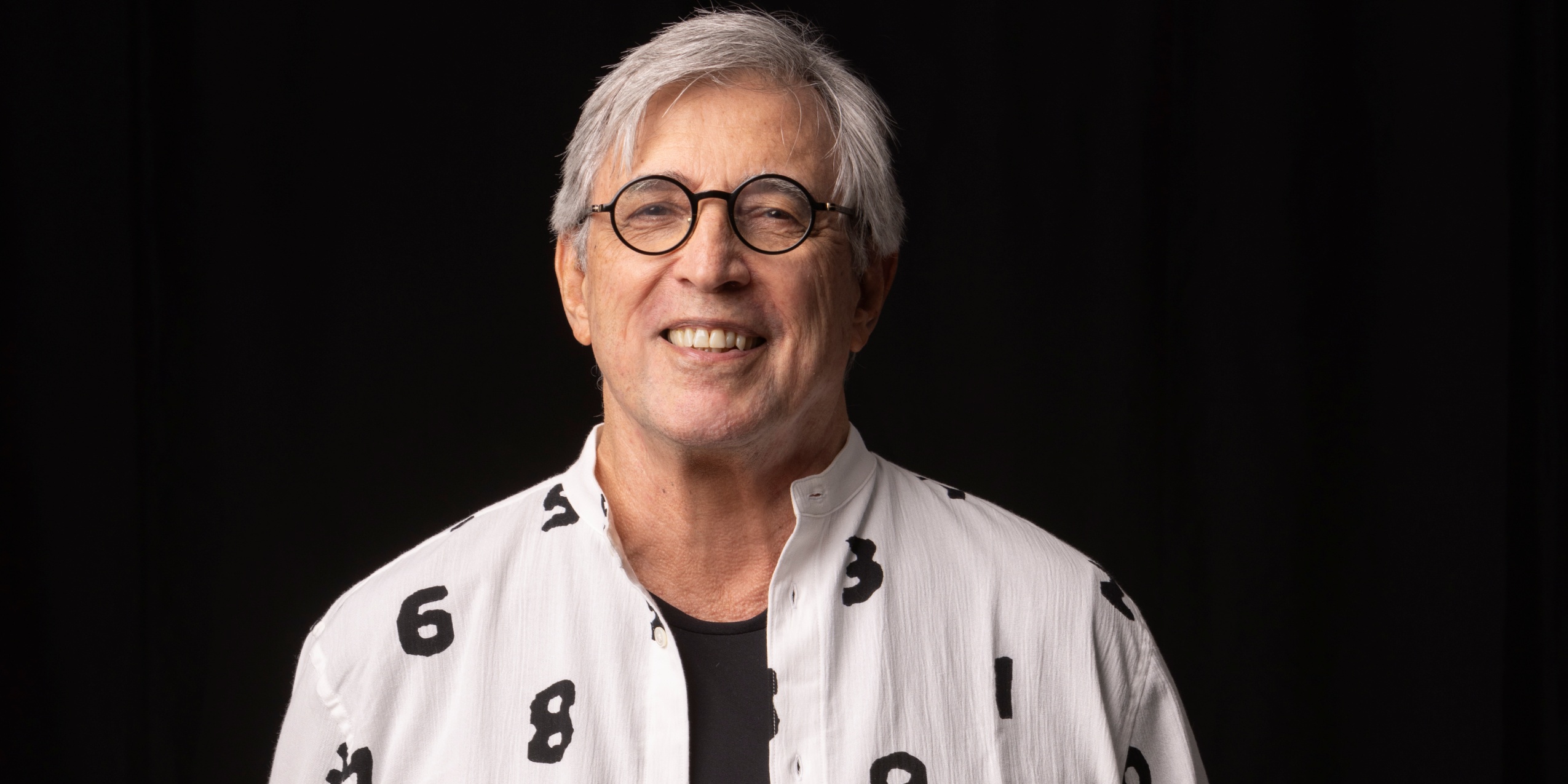 Mand med briller i hvid skjorte med print, foran sort baggrund.