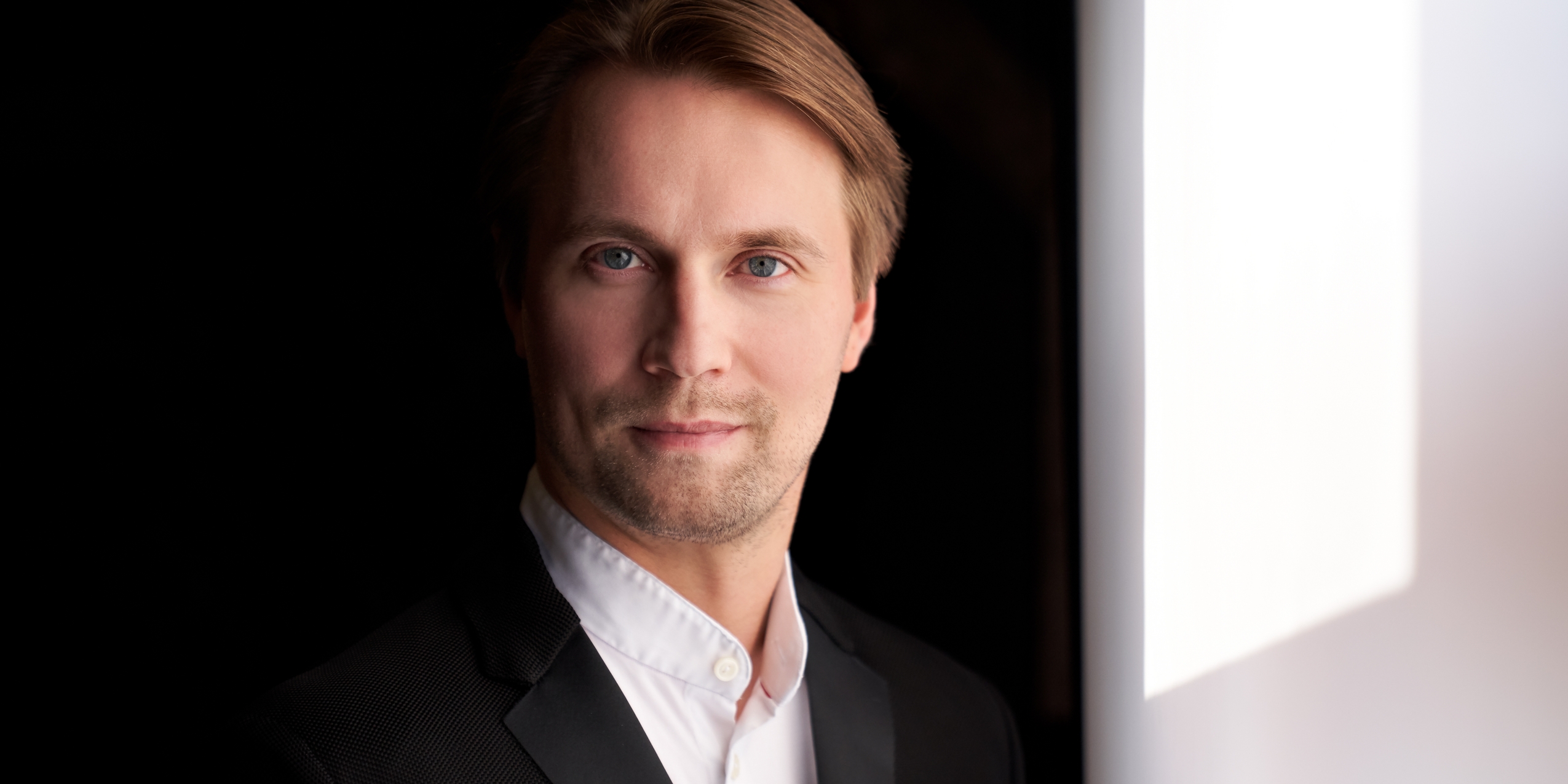 Dirigenten Pietari Inkinen med blå øjne iført hvid skjorte og sort jakke smiler mildt foran sort baggrund