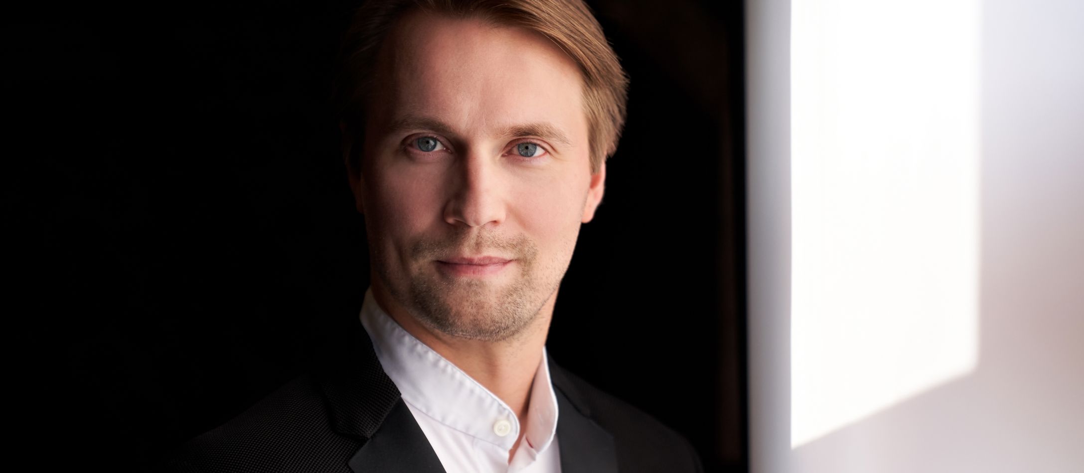 Dirigenten Pietari Inkinen med blå øjne iført hvid skjorte og sort jakke smiler mildt foran sort baggrund