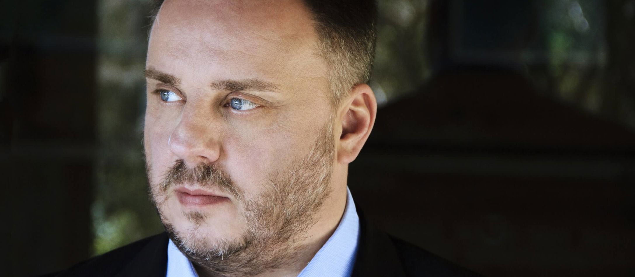 Matthias Goerne kigger med sine blå øjne mod sin højre iført lyseblå skjorte og sort jakke udover på sløret baggrund.