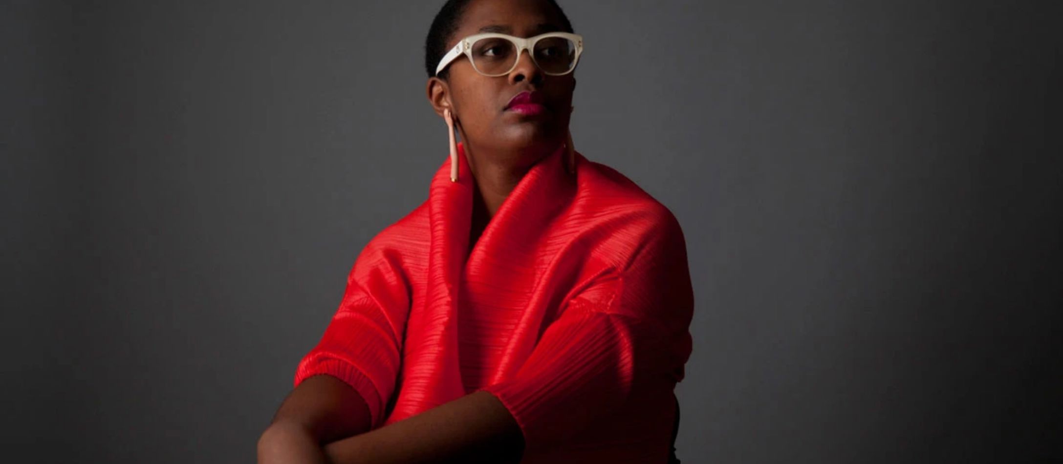 Kvinde i rødt med hvide briller, sidder foran grå baggrund.