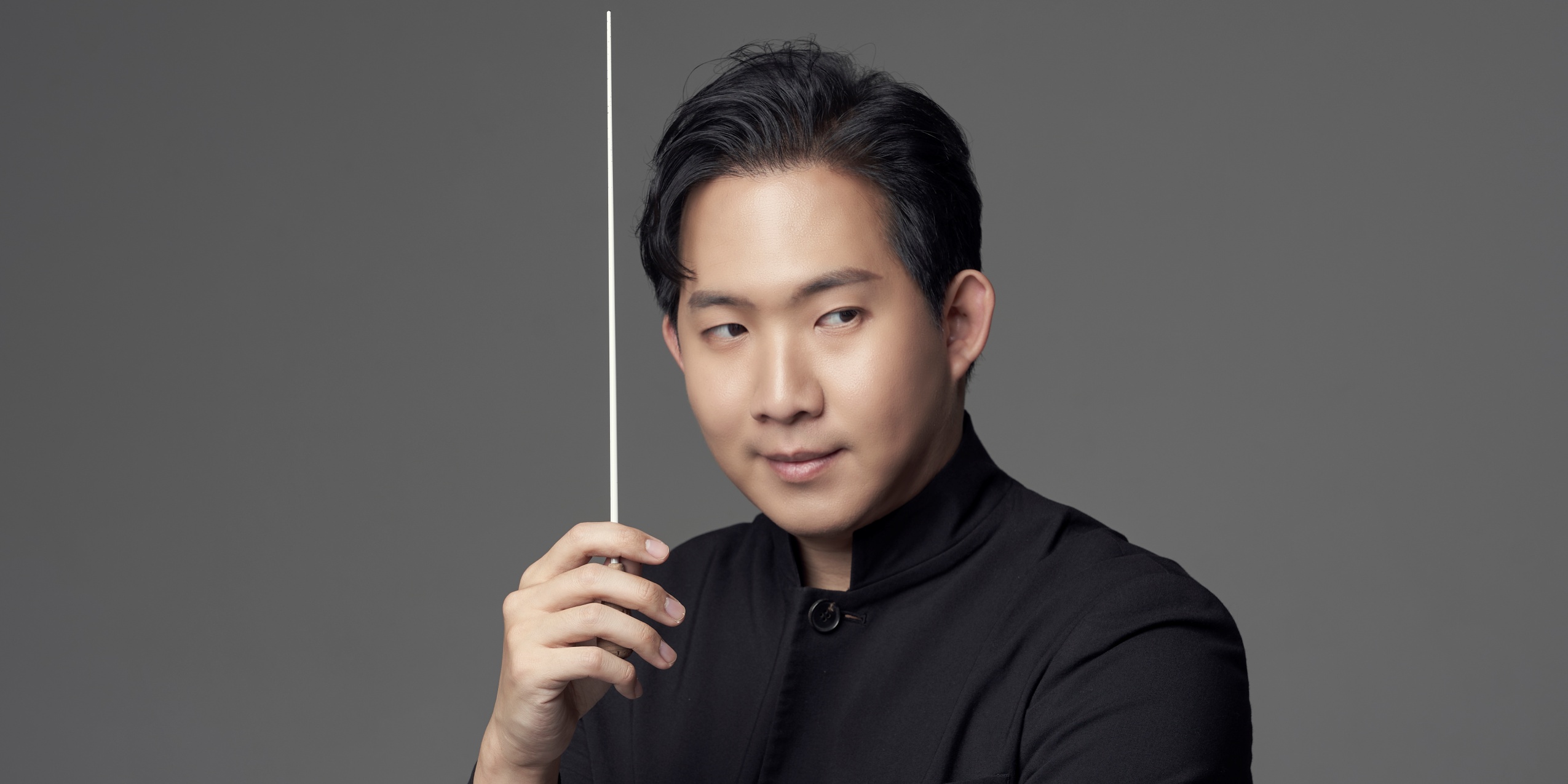 Mand i sort med en dirigentstok foran grå væg.