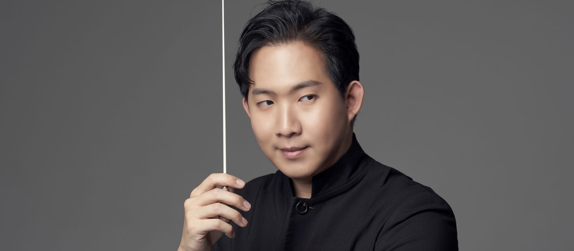 Mand i sort med en dirigentstok foran grå væg.